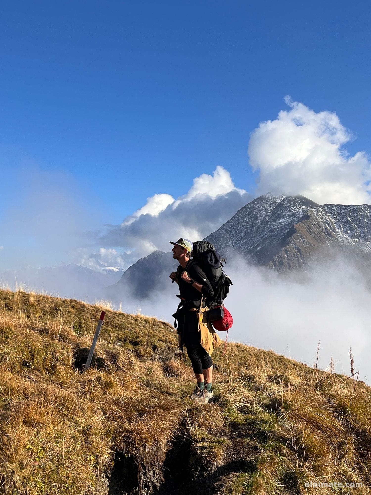 Un alpiniste, sac au dos, sur une crête face à un sommet enneigé enveloppé de nuages